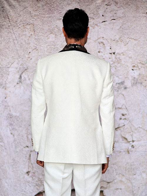 A man in an Insomnia MANZINI GIORGIO 3PC Slim Fit Fancy Floral Pattern White Tuxedo with a bead lapel stands against a textured light-colored wall.