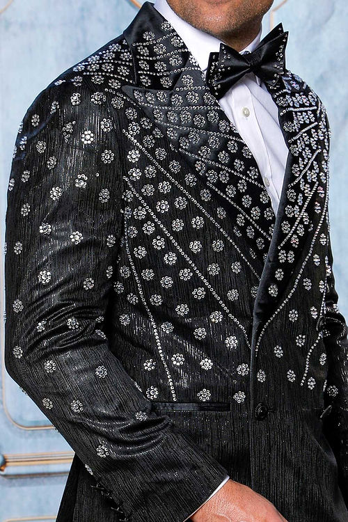 A person in the Insomnia MANZINI GQ-400 Slim Fit Rhinestone Double Breasted Black Tuxedo stands against an ornate blue background, the satin catching light elegantly.