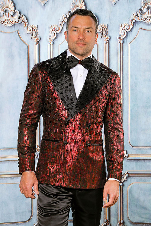 A man in an Insomnia MANZINI GQ-400 2-piece slim fit rhinestone double breast red tuxedo jacket with a bow tie stands against a decorative wall.