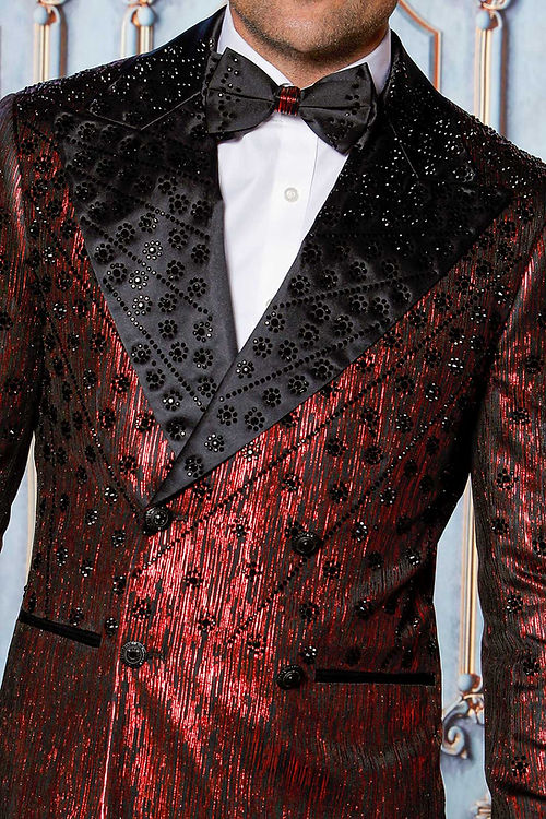 A man in an Insomnia MANZINI GQ-400 2-piece slim fit rhinestone double breast red tuxedo jacket with a bow tie stands against a decorative wall.