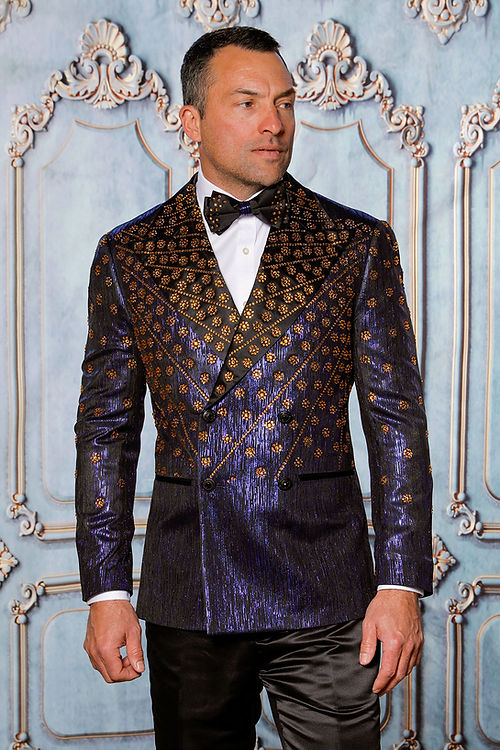 A man wearing the Insomnia MANZINI GQ-400 Rhinestone Slim Fit Royal Tuxedo Jacket, featuring a black and gold pattern with a bow tie, stands before an ornate decorative wall.