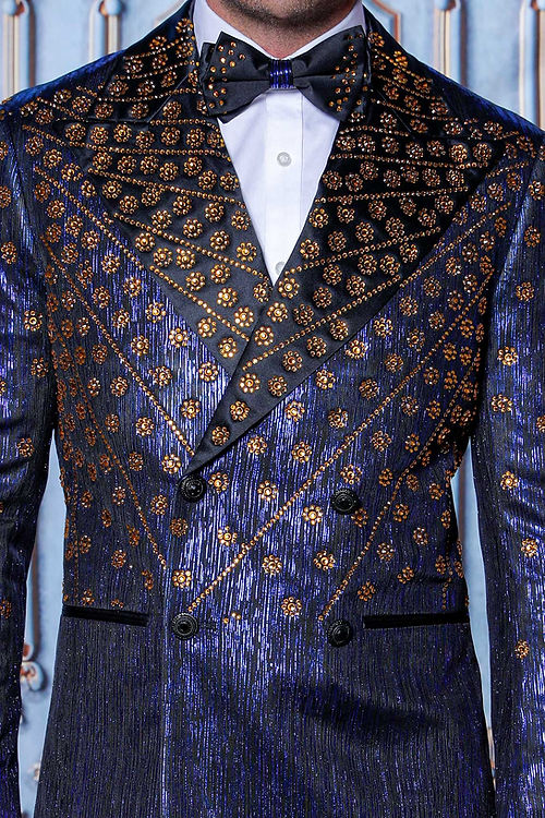A man wearing the Insomnia MANZINI GQ-400 Rhinestone Slim Fit Royal Tuxedo Jacket, featuring a black and gold pattern with a bow tie, stands before an ornate decorative wall.