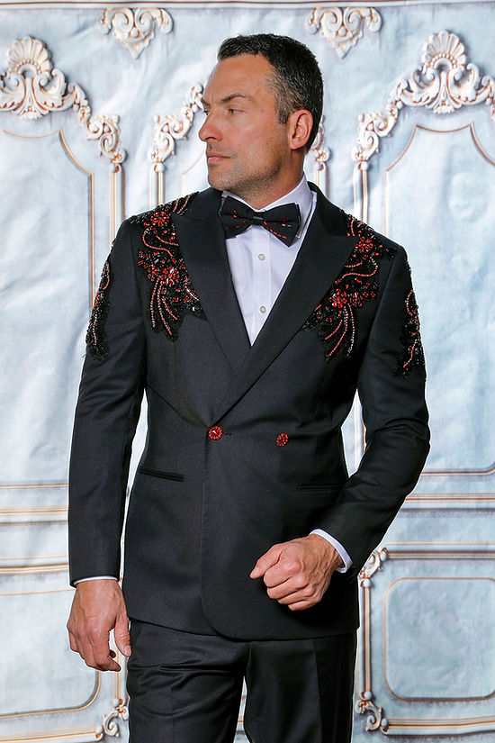 A man in an Insomnia MANZINI GQ300 2-Piece Slim Fit Rhinestone Pattern Double Breast Black/Red Tuxedo Suit and bow tie stands against an ornate floral backdrop, looking to the side.