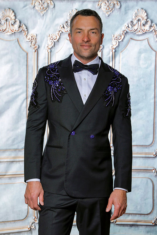A man wears the Insomnia MANZINI GQ300 slim fit double-breasted black/royal tuxedo suit with rhinestone pattern, standing in front of an ornate, light-colored backdrop.