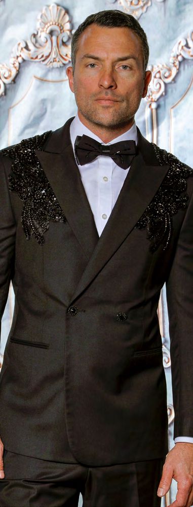 A man in an Insomnia MANZINI GQ300 Slim Fit Rhinestone Pattern Double Breast Black Tuxedo Suit, paired with a floral bow tie, stands against an ornate background.