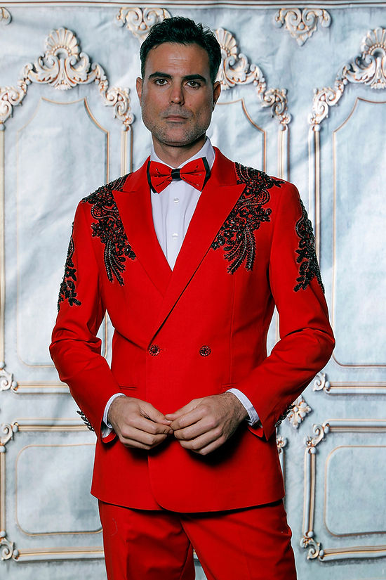 A man wears the Manzini GQ300 2-piece slim fit rhinestone pattern double-breast red tuxedo suit, standing before an ornate backdrop.