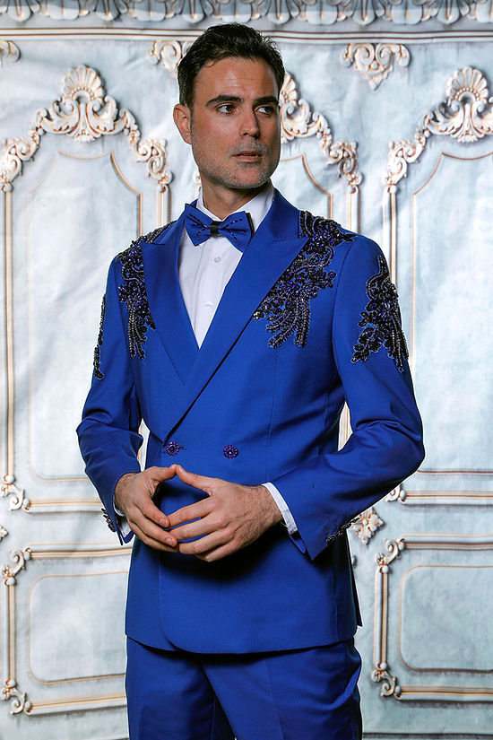 A man wears the Insomnia MANZINI GQ300 2-piece royal blue rhinestone pattern slim fit double-breast tuxedo suit, standing before an ornate patterned background. Sizes: 42R–50R. Final sale, in-store only.