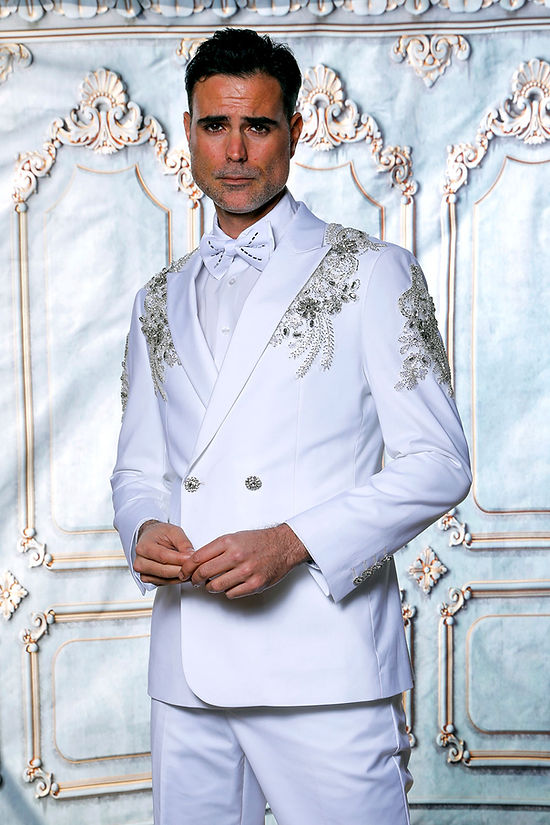 A man in an Insomnia MANZINI GQ300 2-Piece Slim Fit Rhinestone Pattern Double Breast White Tuxedo Suit stands against a decorative backdrop.