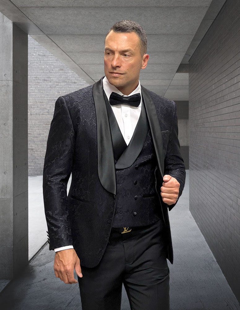 A man in the STATEMENT CLOTHING | GRAMMY-BLACK velvet tuxedo by Statement Clothing, featuring a modern fit, bow tie, and patterned hunter-colored jacket, stands in a sleek concrete hallway, looking to the side.