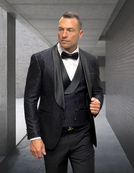 A man in the STATEMENT CLOTHING | GRAMMY-BLACK velvet tuxedo by Statement Clothing, featuring a modern fit, bow tie, and patterned hunter-colored jacket, stands in a sleek concrete hallway, looking to the side.