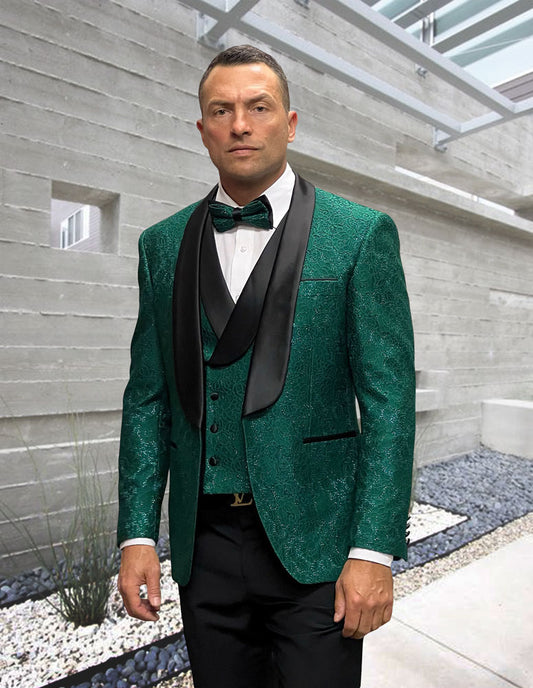Wearing the STATEMENT CLOTHING | GRAMMY-HUNTER by Statement Clothing, a man in a green patterned tuxedo jacket with black lapels, white shirt, bow tie, and solid pants stands amid modern concrete and rock landscaping.