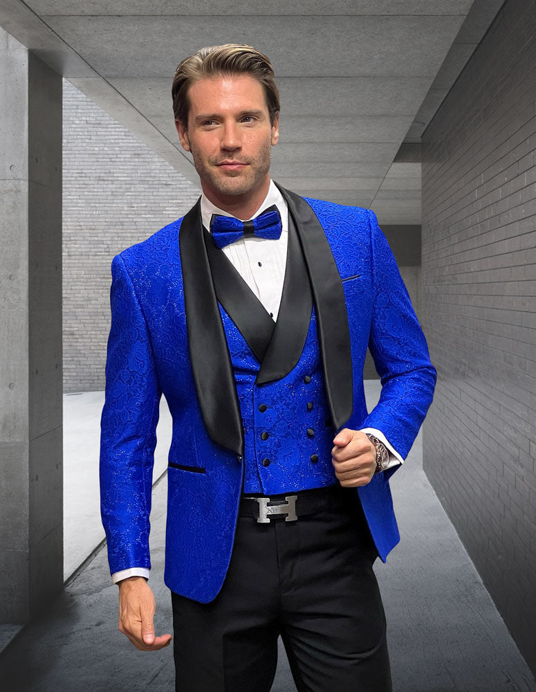 A man stands in a modern concrete corridor wearing the STATEMENT CLOTHING | GRAMMY-SAPPHIRE tuxedo by Statement Clothing—a fancy blue patterned jacket with black lapels, double-breasted vest, white shirt, blue bow tie, and solid black pants.