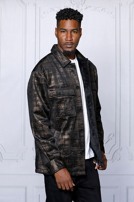 A man in a MANZINI GREECE gold metallic fabric button-down coat by Insomnia stands against a light-colored, ornate backdrop, exuding sophistication.