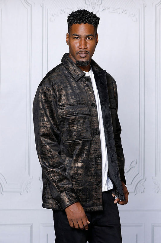 A man in a MANZINI GREECE gold metallic fabric button-down coat by Insomnia stands against a light-colored, ornate backdrop, exuding sophistication.