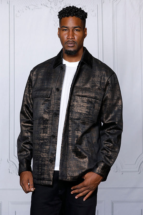 A man in a MANZINI GREECE gold metallic fabric button-down coat by Insomnia stands against a light-colored, ornate backdrop, exuding sophistication.