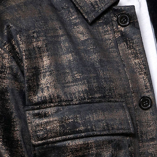 A man in a MANZINI GREECE gold metallic fabric button-down coat by Insomnia stands against a light-colored, ornate backdrop, exuding sophistication.