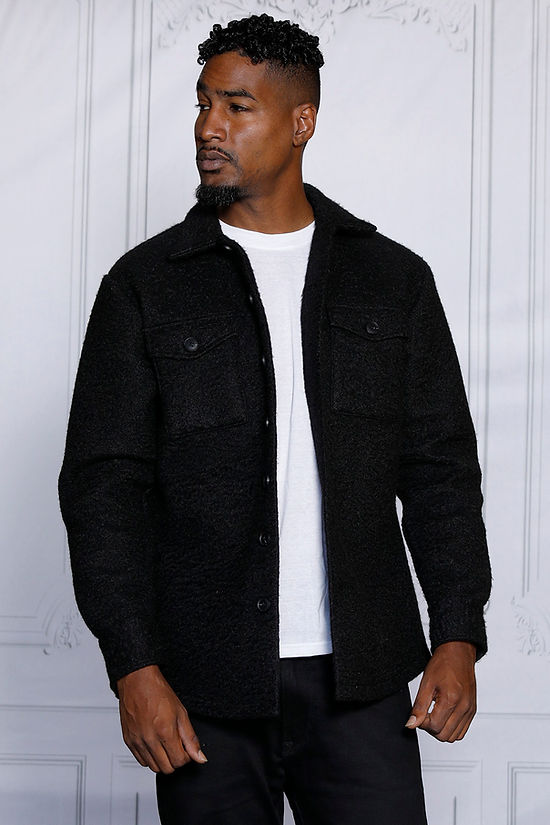 A man stands by a decorative white wall, gazing right, wearing an Insomnia MANZINI ICELAND Black Soft Fleece Button Sweater Shirt over a white shirt. This super soft poly fleece piece adds effortless style to his look.