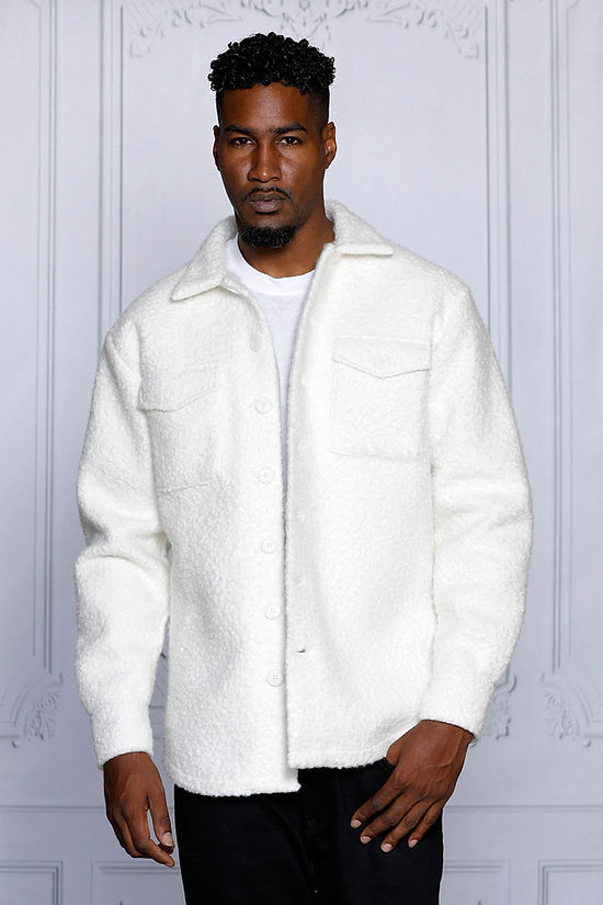 A man, exuding casual sophistication, stands against a light grey decorative wall wearing an Insomnia MANZINI ICELAND White Soft Fleece Button Sweater Shirt.