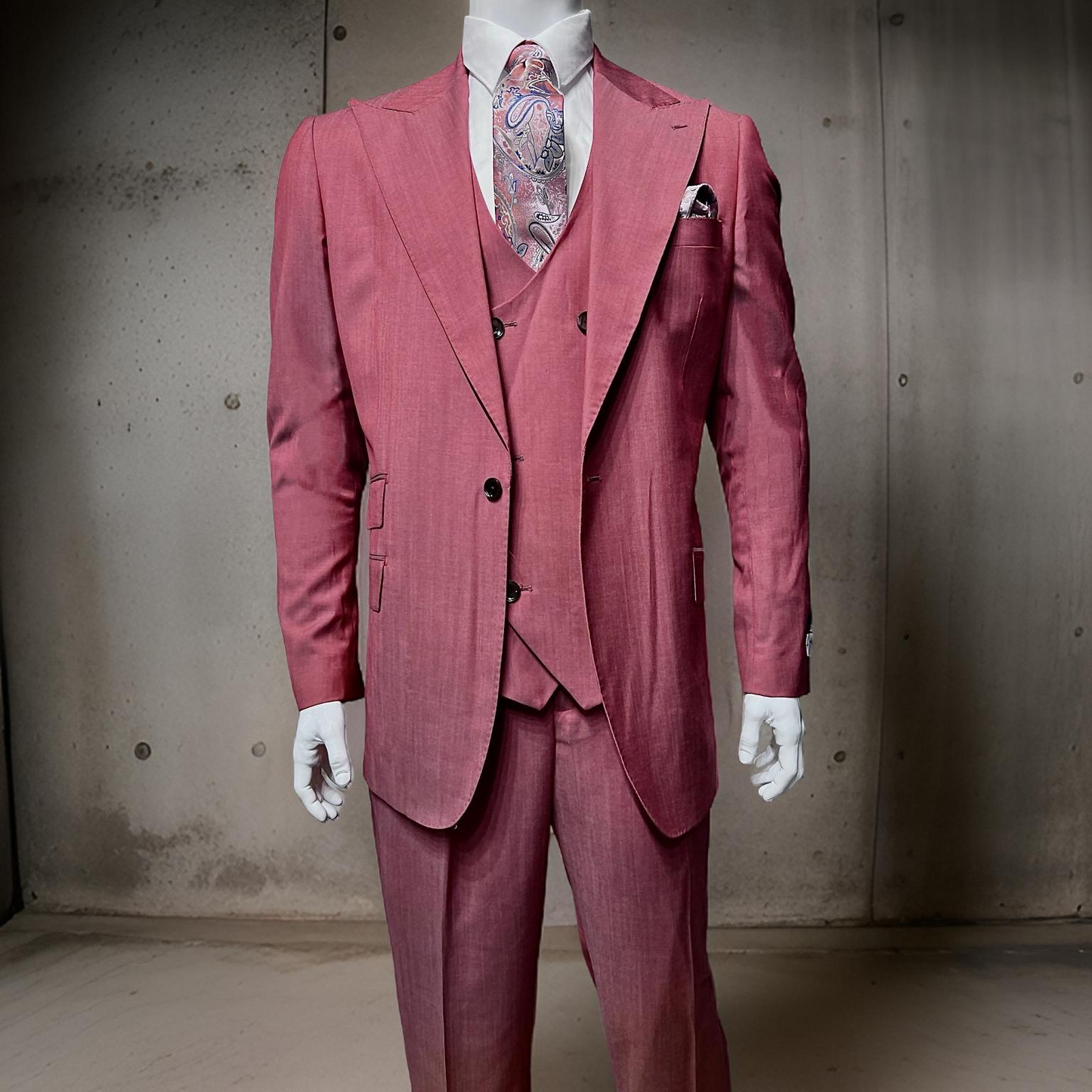 The mannequin features the Tiglio Rosso Orvietto Light Red TL4016, a wool three-piece suit in pink (sizes 48R, 50R, 52R), accompanied by a paisley tie and pocket square. Its elegance contrasts with the plain concrete backdrop, illustrating Tiglio Rosso's timeless style.