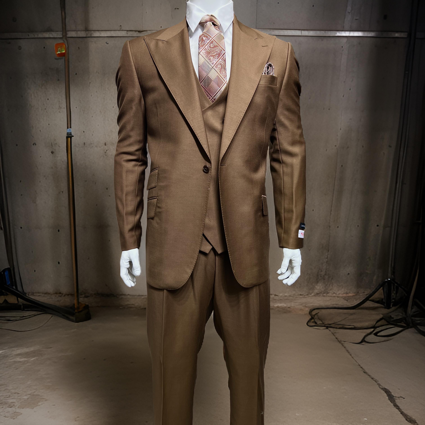 In a concrete-walled room, a mannequin is attired in a Tiglio Rosso Orvietto Solid Tobacco single-pleat three-piece suit paired with a patterned tie.