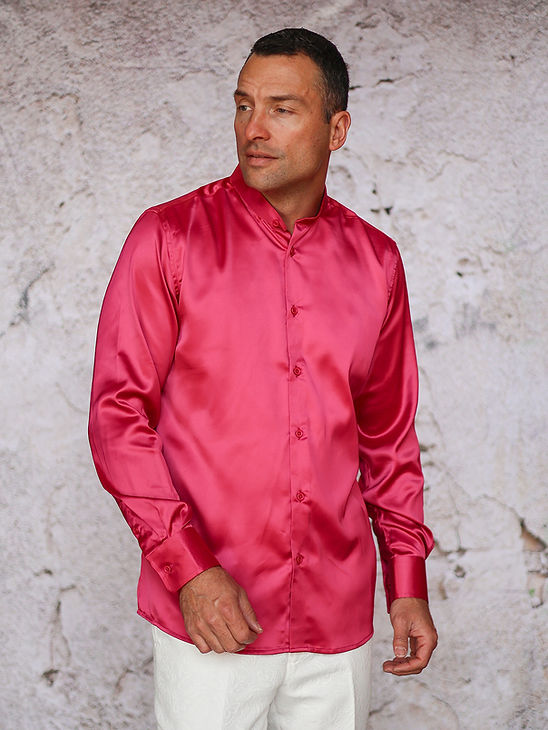 A man wears the Manzini IN-101 Regular Collar Solid Color Satin Shirt in fuchsia and white pants, standing before a textured light wall.