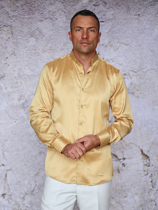A man stands against a textured light wall, wearing the Manzini IN-101 Regular Collar Solid Color Satin Shirt in gold and white pants, looking directly at the camera with hands clasped.