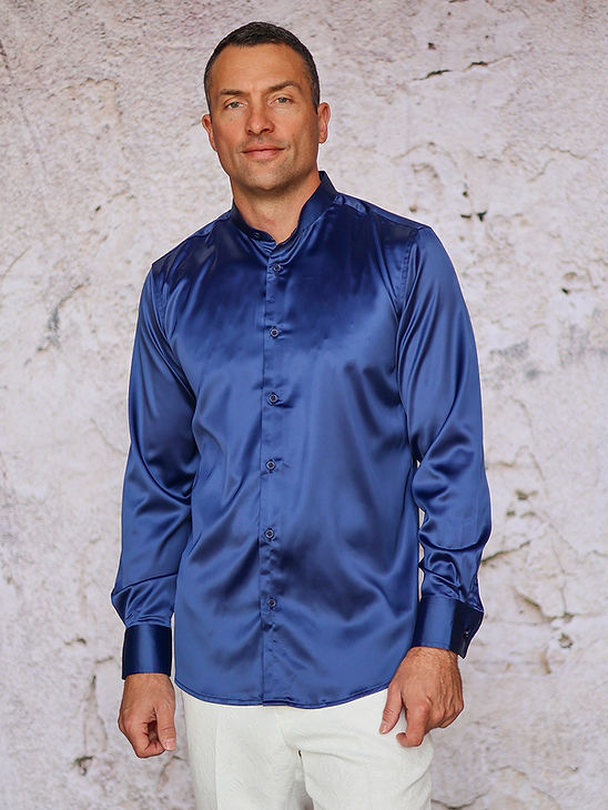 A man wears the Manzini IN-101 Regular Collar Solid Color Satin Shirt in navy by Manzini with white pants, standing before a light textured wall, ready for formal events.