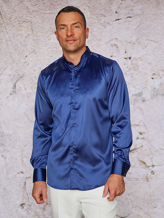 A man wears the Manzini IN-101 Regular Collar Solid Color Satin Shirt in navy by Manzini with white pants, standing before a light textured wall, ready for formal events.