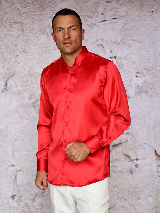 A man wears the MANZINI IN-101 Regular Collar Solid Color Satin Shirt in red by Manzini and white pants, standing before a textured light-colored wall.