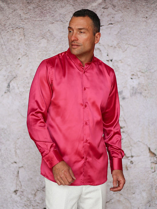 A man wears the Manzini INR-102 Banded Collar Solid Color Satin Shirt in fuchsia with white pants, standing before a textured light-colored wall.
