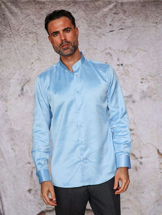 A man wears the Manzini INR-102 Banded Collar Solid Color Satin Shirt in Sky with dark pants, standing before a textured, light backdrop.