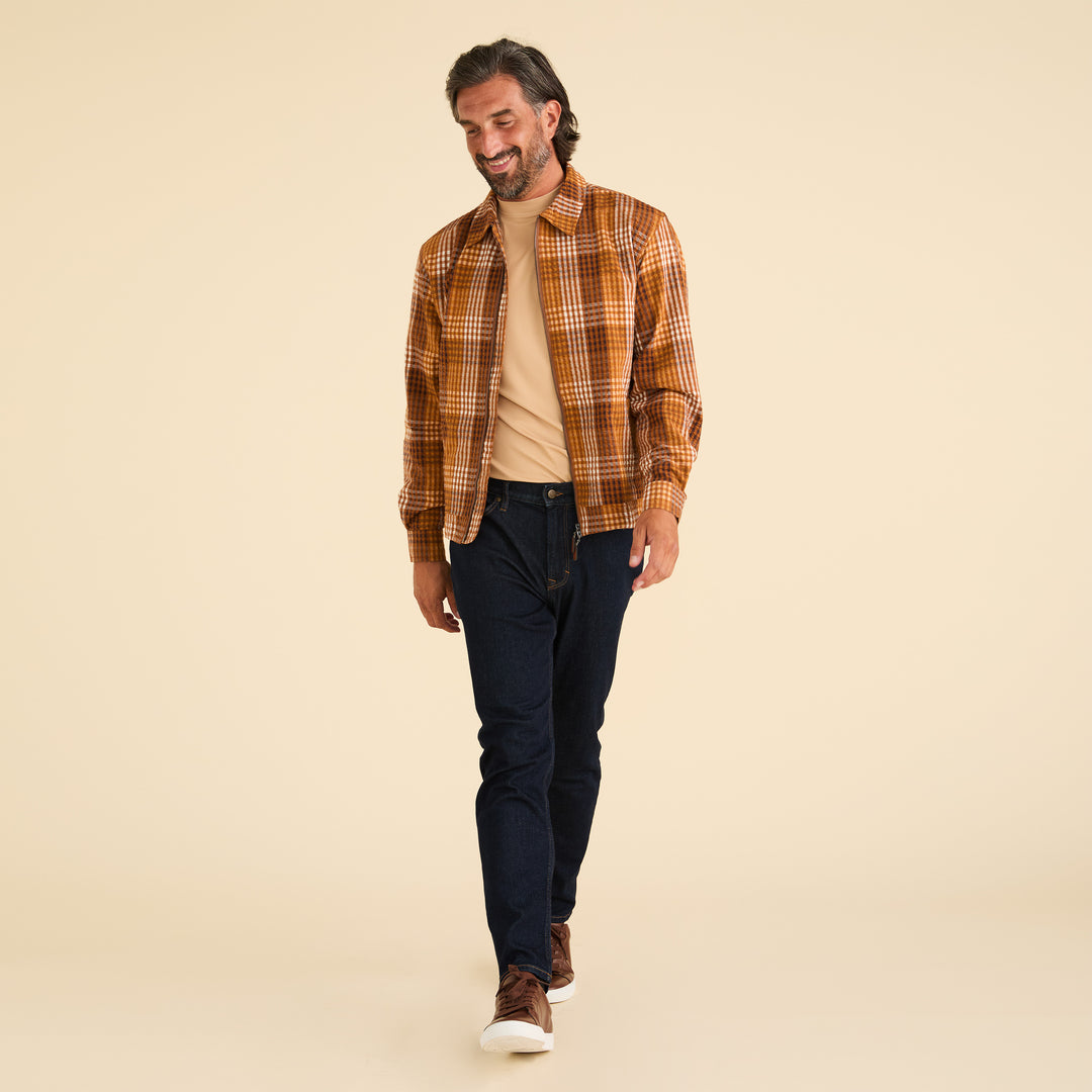 A man strides confidently, sporting a brown plaid shirt over a beige t-shirt, dark jeans, and brown sneakers. His subtle elegance is reminiscent of Inserch's Seersucker Gingham Jacket JS272-00027 Aztec against the neutral backdrop.