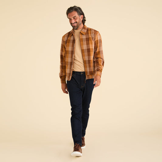 A man strides confidently, sporting a brown plaid shirt over a beige t-shirt, dark jeans, and brown sneakers. His subtle elegance is reminiscent of Inserch's Seersucker Gingham Jacket JS272-00027 Aztec against the neutral backdrop.