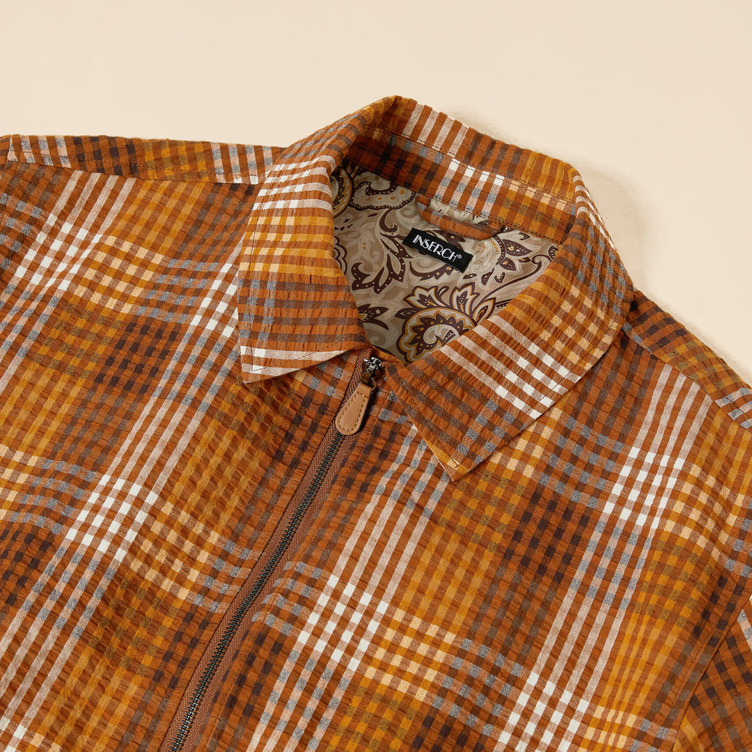 A man strides confidently, sporting a brown plaid shirt over a beige t-shirt, dark jeans, and brown sneakers. His subtle elegance is reminiscent of Inserch's Seersucker Gingham Jacket JS272-00027 Aztec against the neutral backdrop.