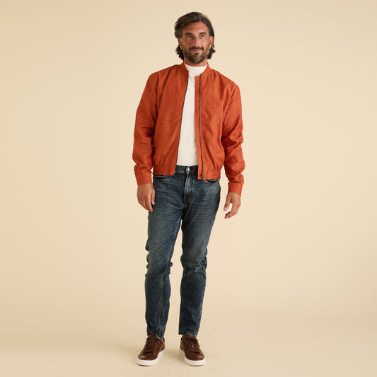 A man faces forward against a beige background, wearing the Inserch Linen Bomber Jacket JS660-00109 in burnt orange, paired with a white turtleneck, blue jeans, and brown sneakers.