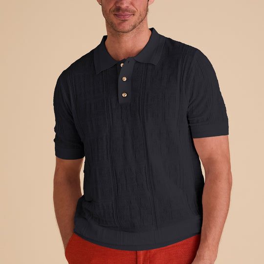 A man wears the Inserch SS Knit Polo KS001-00001 Black, a textured geometric knit short-sleeve shirt with novelty buttons by Inserch, paired with red pants against a beige background.