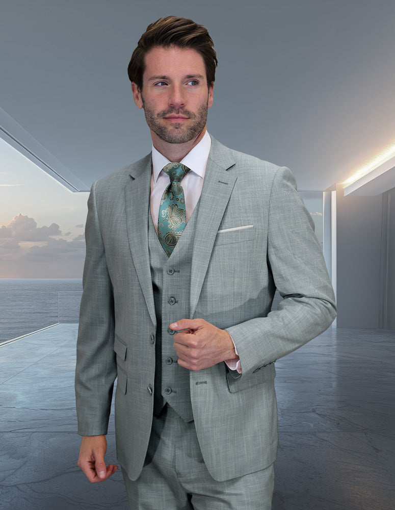 A man in the STATEMENT CLOTHING | LARZO-AQUA modern fit sharkskin Italian wool suit by Statement Clothing, paired with a patterned aqua green tie, stands in a sleek room with large windows overlooking the ocean.