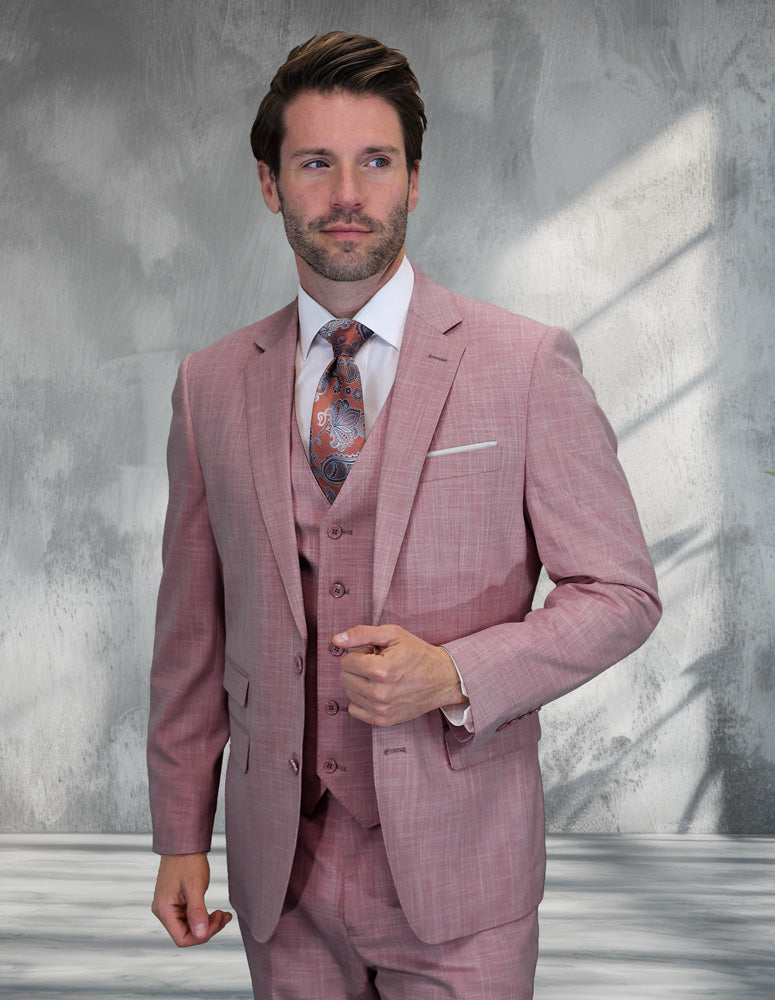 A man wearing the STATEMENT CLOTHING | LARZO-BLUSH modern fit Italian wool suit by Statement Clothing, paired with a white shirt and patterned tie, stands before a textured gray wall with sunlight streaming in.