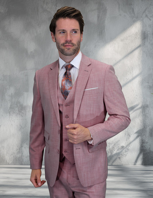 A man wearing the STATEMENT CLOTHING | LARZO-BLUSH modern fit Italian wool suit by Statement Clothing, paired with a white shirt and patterned tie, stands before a textured gray wall with sunlight streaming in.