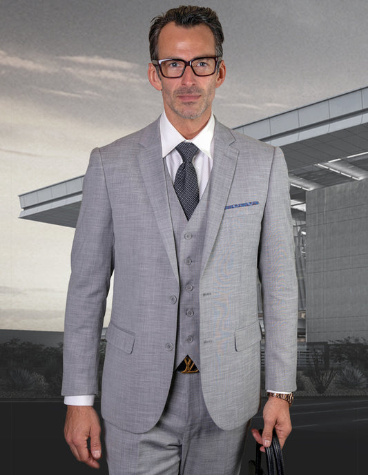A man wearing the STATEMENT CLOTHING | LAZARO-GREY slim fit three-piece suit by Statement Clothing, paired with a white shirt, patterned tie, and glasses, stands before a modern building holding a black bag.