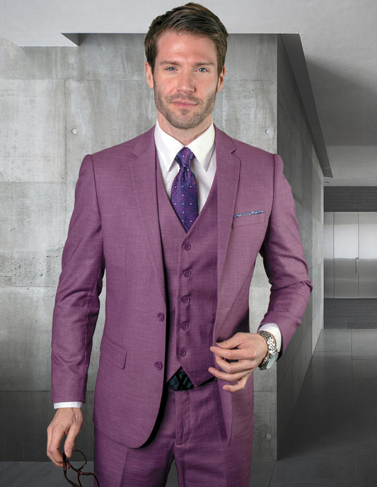 A man in a slim fit STATEMENT CLOTHING | LAZARO-MAUVE three-piece suit by Statement Clothing stands indoors with eyeglasses in hand, while modern concrete walls accentuate the elegance of his Italian fabric ensemble.
