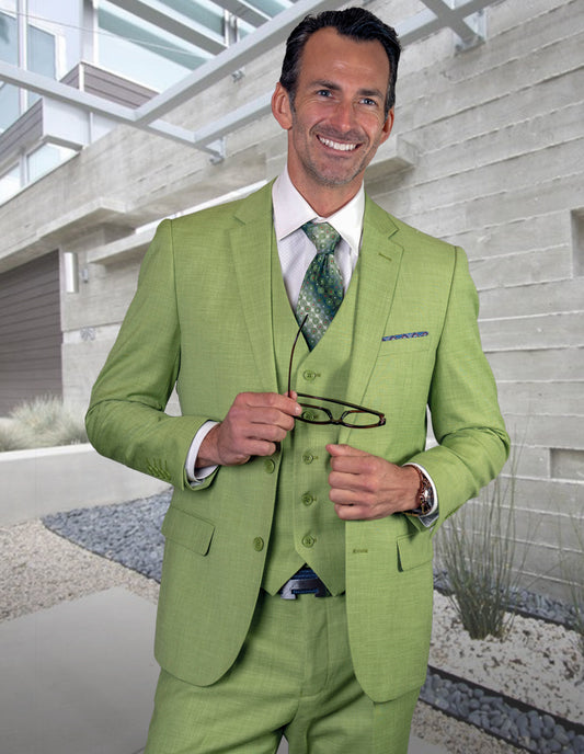 A man wears the STATEMENT CLOTHING | LAZARO-PISTACHIO slim fit suit by Statement Clothing, crafted from fine Italian fabric, as he stands outside a modern building, smiling at the camera and holding eyeglasses.