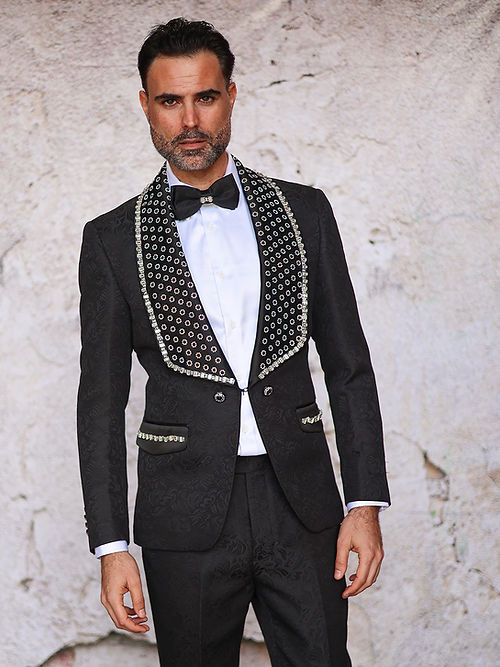 A person wearing the Insomnia MANZINI LEONARDO 2-Piece Fancy Rhinestone Black Tuxedo Suit stands against a textured white wall, with the rhinestones subtly catching the light.