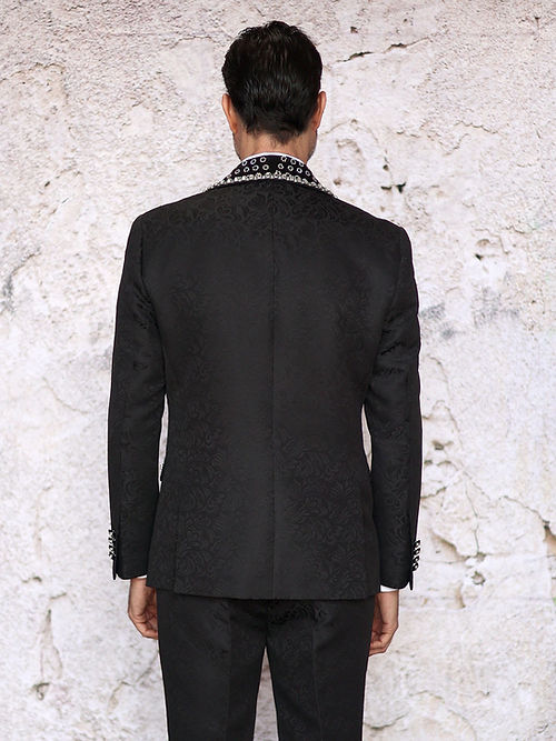 A man wears an Insomnia MANZINI LEONARDO 2-piece fancy rhinestone black tuxedo suit with a patterned lapel and bow tie, crafted from shiny fabric, standing against a textured background.