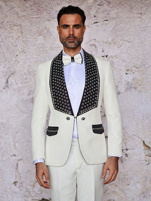 A man in an Insomnia MANZINI LEONARDO 2-Piece Fancy Rhinestone Cream Tuxedo Suit featuring a circle-patterned shawl collar and matching bow tie stands against a textured wall.