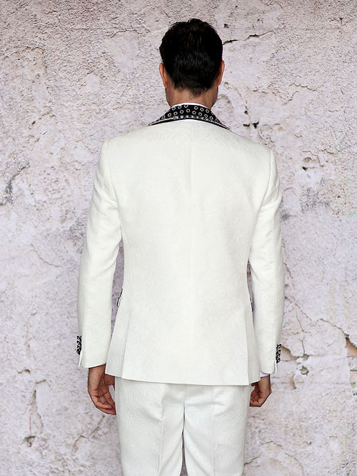 A man in an Insomnia MANZINI LEONARDO 2-Piece Fancy Rhinestone Cream Tuxedo Suit featuring a circle-patterned shawl collar and matching bow tie stands against a textured wall.