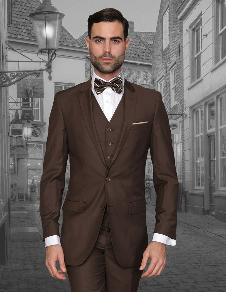 A man in the Statement Clothing LORENZO-BROWN slim fit Italian wool three-piece suit with a bow tie stands on a cobblestone street with historic buildings behind him.