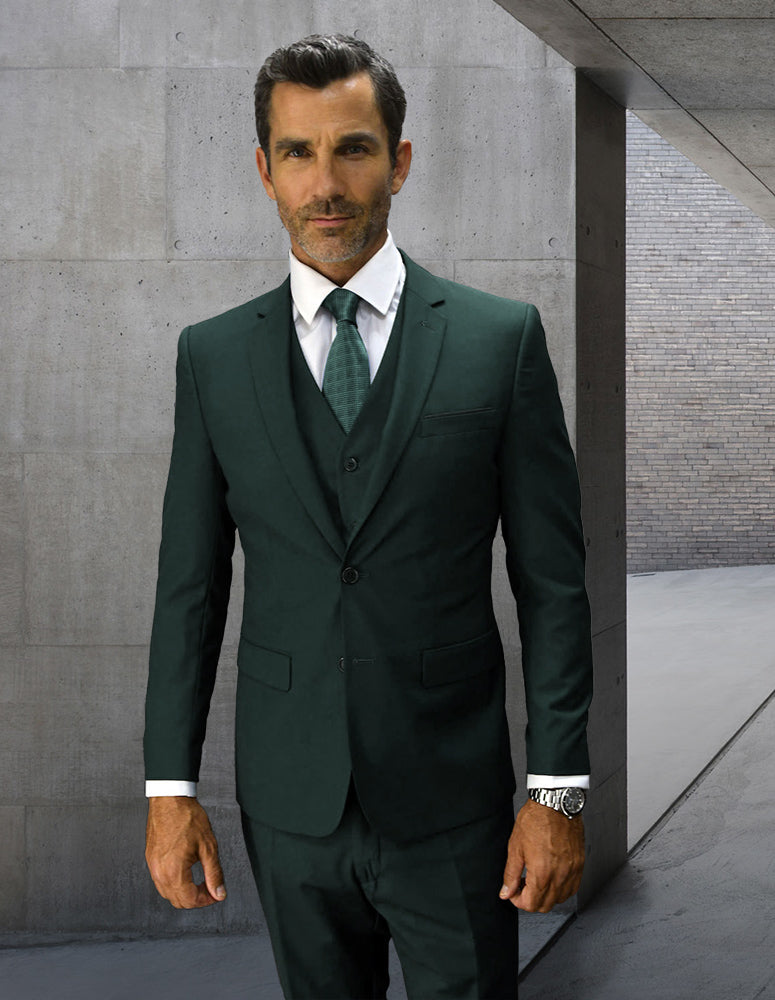 A man in the STATEMENT CLOTHING | LORENZO-HUNTER dark green Italian wool suit by Statement Clothing, paired with a white shirt and green tie, stands in a modern concrete corridor with a neutral expression.