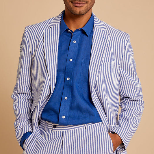 A man wears the Inserch Linen Long-Sleeve Shirt LS29116-00190 in Estate Blue, styled with a white suit featuring thin blue stripes, one hand in his pocket, set against a beige background.
