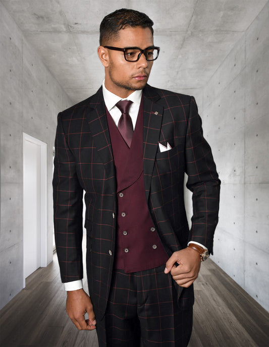 A man in the STATEMENT CLOTHING | LUCCA-BLACK-BURGUNDY modern fit Italian wool plaid suit with maroon vest and tie, by Statement Clothing, stands in a sleek hallway with concrete walls and wooden floors.